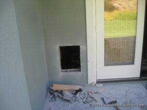 Hole for pet door through stucco from the outside, next to a sliding glass door. The door has not yet been installed. There is debris from the stucco below the pet door.