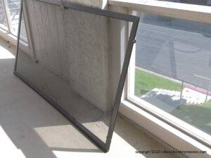 Black-framed screen door on its side on the ground propped up against a window pane.